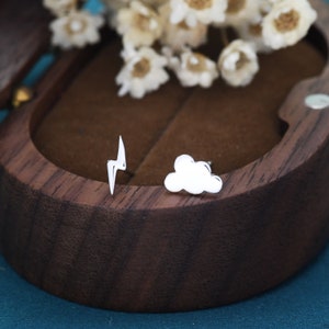 May include: A pair of silver earrings, one shaped like a cloud and the other shaped like a lightning bolt. The earrings are displayed in a wooden box.