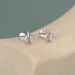 May include: A pair of silver-toned stud earrings shaped like small airplanes. Each earring features a polished metallic finish with a flat back post and a visible 925 stamp on the circular base, designed for secure placement in a piercing.