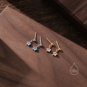 Op de afbeelding: Twee paar stervormige oorbellen met iriserende blauwe stenen. Het ene paar is zilver en het andere is goud.