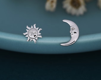 May include: A pair of silver stud earrings. One earring is a sun with a face, and the other is a crescent moon with a face. The earrings are on a teal surface.