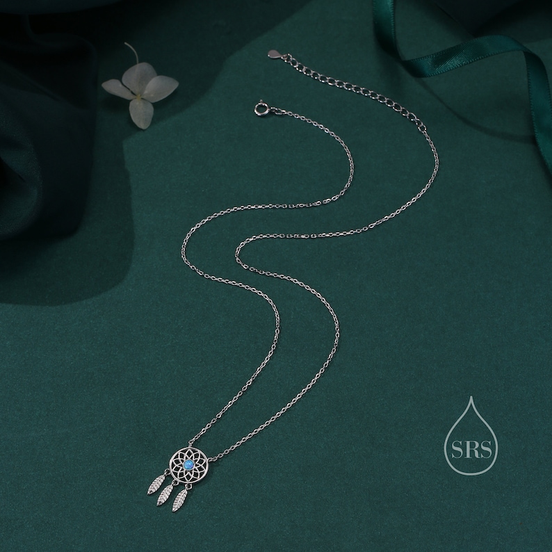May include: A silver-toned necklace featuring a circular dreamcatcher pendant with a small, round, light blue stone at its center. Three delicate silver-toned feather charms hang from the bottom of the pendant. The chain includes a lobster clasp.