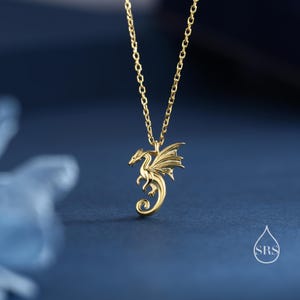 May include: A gold-toned necklace featuring a delicate chain and a pendant shaped like a small dragon with detailed wings and a curled tail. The dragon is depicted in a side profile, suspended from the chain, with a small watermark logo reading SRS.