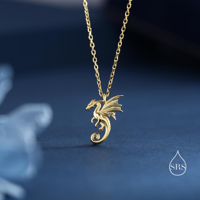 May include: A gold-toned necklace featuring a delicate chain and a pendant shaped like a small dragon with detailed wings and a curled tail. The dragon is depicted in a side profile, suspended from the chain, with a small watermark logo reading SRS.