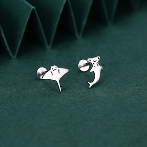 May include: A pair of silver stud earrings. One earring is shaped like a manta ray, the other like a hammerhead shark. Both earrings have a post and backing for secure wear. The earrings are set against a dark green background.
