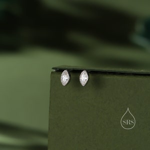 May include: A pair of silver earrings with clear marquise-shaped stones. The earrings are on a green surface.