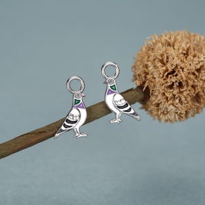 May include: Two silver-toned metal charms shaped like pigeons, each featuring enamel detailing in shades of green, purple, and black on the neck and wings. Each charm includes a circular jump ring at the top for attachment to jewelry or accessories.