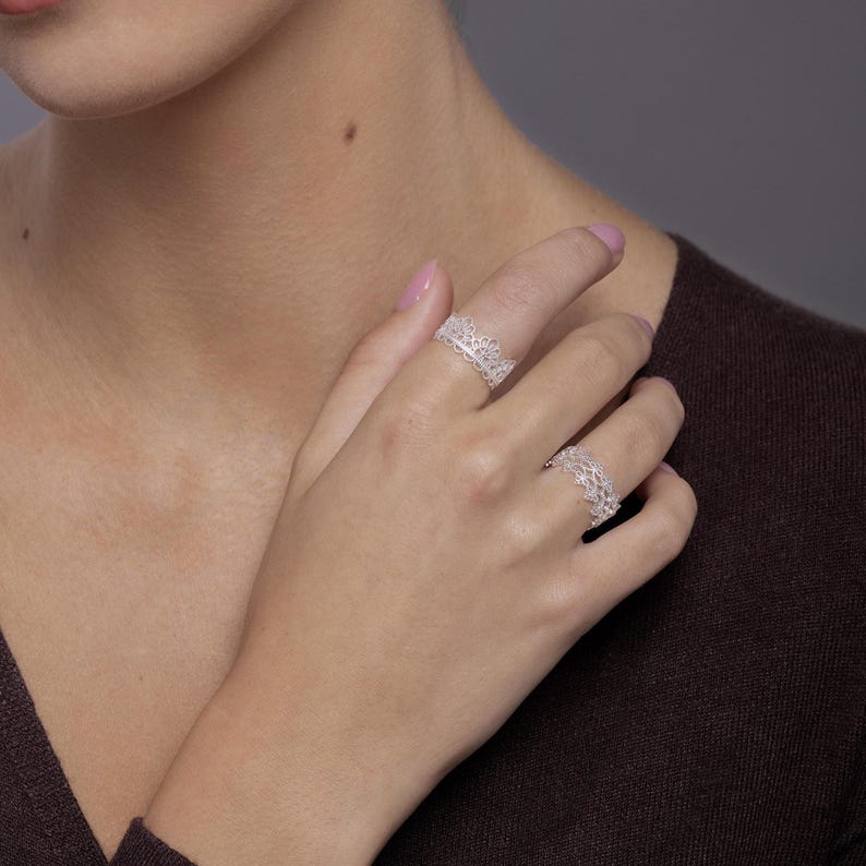 May include: Two silver rings featuring an intricate, openwork lace-like pattern. The rings are worn on the index and ring fingers, showcasing a delicate, filigree-style metal design that wraps around the finger with a scalloped edge.