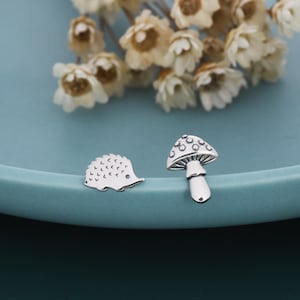 May include: Two small, silver charms: a hedgehog and a mushroom. The hedgehog charm has a spiky back and a small eye. The mushroom charm has a dotted cap and a stem. The charms are on a blue surface, with white flowers in the background.