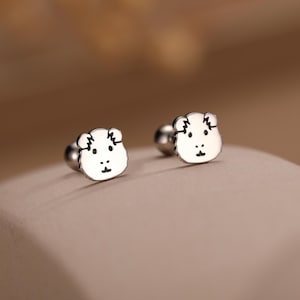 Könnte beinhalten: Ein Paar silberfarbene Ohrstecker. Jeder Ohrring zeigt ein kleines, weißes Bärengesicht mit schwarzen Details für Augen, Nase und Mund. Die Ohrringe haben einen Stecker und Kugelverschluss.