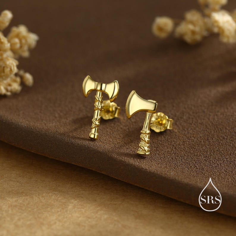 May include: A pair of gold-colored axe-shaped stud earrings. The earrings have detailed handles and axe heads. The earrings are displayed on a brown surface, with a blurred background.