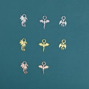 Pode incluir: Nove pingentes de dragão de metal em prata, ouro e ouro rosa. Os pingentes apresentam desenhos detalhados de dragões com asas e caudas enroladas. Os pingentes estão dispostos sobre um fundo verde-azulado.