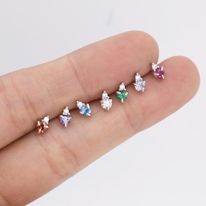 May include: A set of six small, silver stud earrings with a single, round gemstone in the center and two smaller, round, clear gemstones on the side. The gemstones are in the colors red, purple, blue, green, pink, and clear.