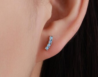 Puede incluir: Un pendiente de plata con cuatro pequeñas piedras de ópalo azul claro dispuestas en línea curva. El pendiente se muestra en una oreja, destacando el diseño delicado y el color sutil de los ópalos.