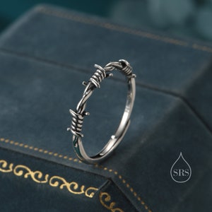 Könnte beinhalten: Ein silberner Ring mit einem Stacheldraht-Design. Der Ring liegt auf einem blauen Samt-Hintergrund.
