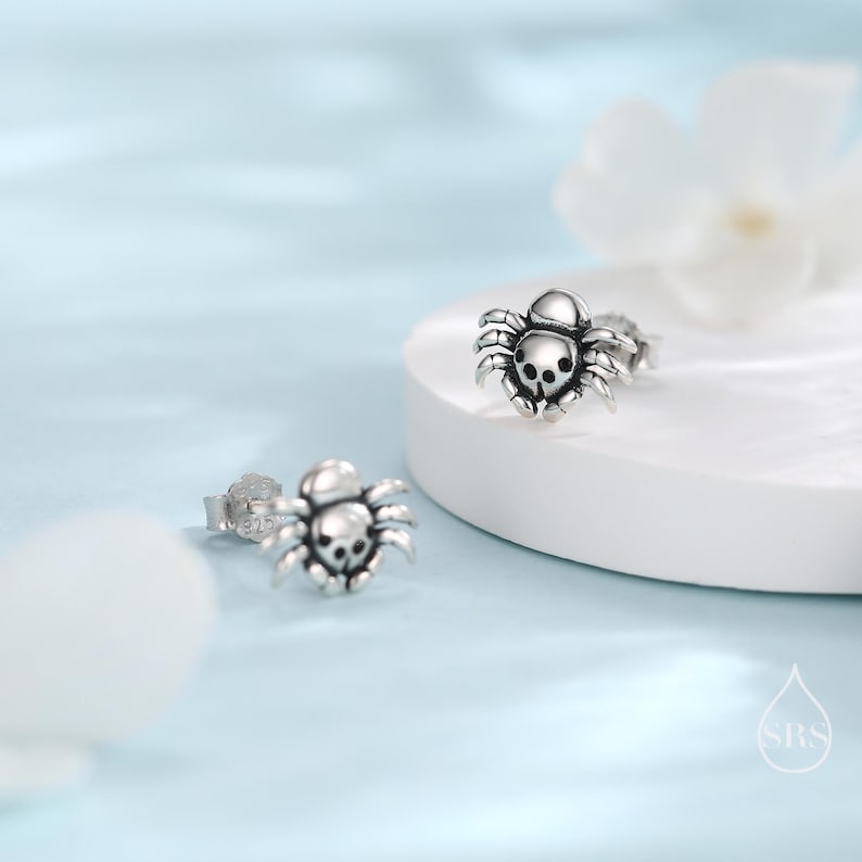 May include: A pair of silver spider earrings. Each earring features a detailed spider design with a skull-like head.