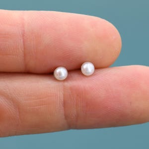 May include: A close-up of two small, round, white pearl earrings. The pearls are set against a blurred background. The earrings are small and delicate, suitable for everyday wear.