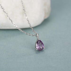 May include: A silver-toned necklace featuring a single pear-shaped purple gemstone pendant. The faceted stone is held in a simple metal setting and hangs from a delicate, thin silver-toned chain with small, uniform oval links.