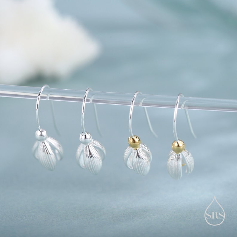 May include: Four silver-toned and gold-toned flower-shaped dangle earrings. Each earring has a small ball at the top and a delicate, petal-like design. The earrings are displayed on a clear rod against a soft blue background.