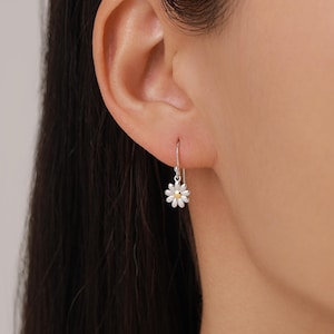 May include: A silver daisy earring with a white petal design and a yellow center. The earring is suspended from a simple hook, showcasing a delicate floral theme. The earring is shown on a person's ear.
