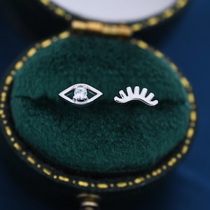 May include: A pair of silver-toned stud earrings. One earring is an eye shape with a clear gemstone center, and the other is a curved eyelash design. The earrings are displayed in a green velvet jewelry box with a gold trim.