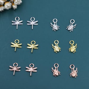 Op de afbeelding: Een verzameling metalen bedels in zilver, goud en roségoud tinten. De bedels bevatten libellen- en keverontwerpen, perfect voor het maken van sieraden of knutselprojecten. De bedels worden gepresenteerd op een tealblauwe achtergrond.