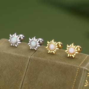 Può includere: Quattro orecchini a bottone con un design a forma di sole. Due sono argentati e due dorati. Ogni orecchino presenta una pietra centrale rotonda di opale bianca. Gli orecchini sono esposti su una superficie di velluto verde.