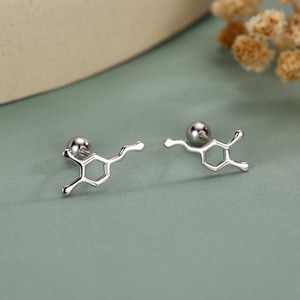 May include: A pair of silver stud earrings, each shaped like a dopamine molecule. The earrings have a small, round stud and a stylized chemical structure. They are presented on a light teal surface.