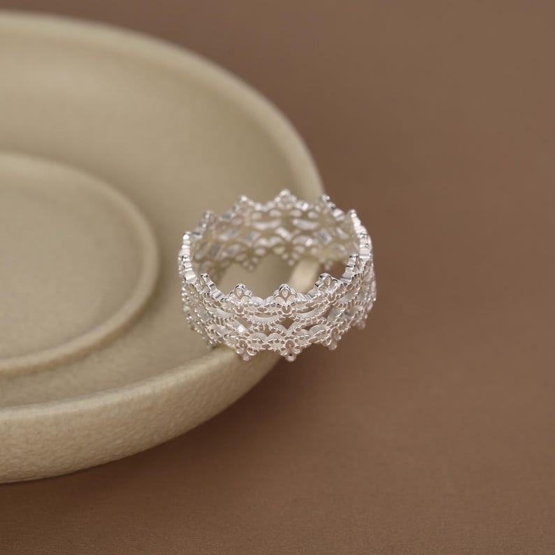 May include: A silver ring featuring an intricate, openwork lace-like pattern with a scalloped edge. The band has a consistent width and a metallic finish, showcasing a detailed filigree design that wraps around the entire circumference of the ring.
