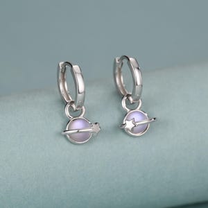 Puede incluir: Un par de pendientes de aro plateados con un colgante en forma de planeta que incluye un anillo y una pequeña estrella en el lateral. El colgante del planeta tiene un centro esférico opaco, liso y de color violeta claro.