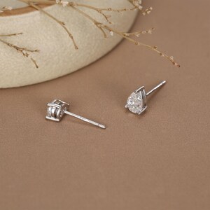 May include: Two silver stud earrings. One earring features a square-cut, clear gemstone. The other earring has a pear-shaped, clear gemstone. Both earrings are set on thin silver posts, displayed on a brown surface.