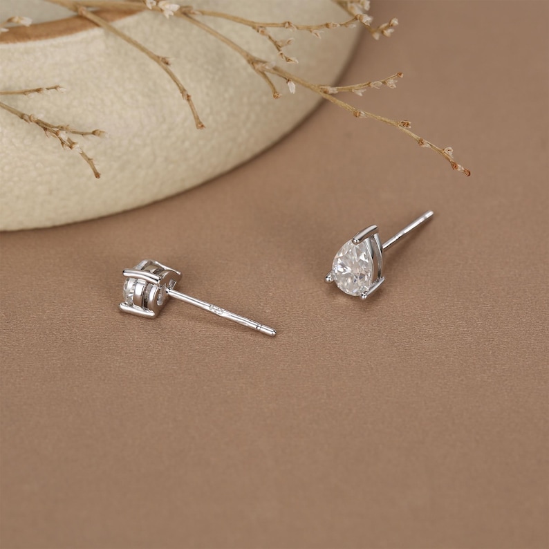 May include: Two silver stud earrings. One earring features a square-cut, clear gemstone. The other earring has a pear-shaped, clear gemstone. Both earrings are set on thin silver posts, displayed on a brown surface.