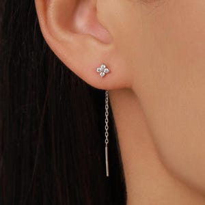 May include: A silver-toned threader earring featuring a four-petaled flower stud design adorned with small clear stones. A delicate chain extends from the back of the flower stud, ending in a thin, straight metal bar that hangs vertically.