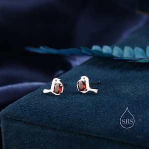 May include: A pair of silver stud earrings featuring a small bird design with a red gemstone in the centre.