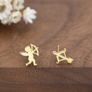 May include: A pair of gold-coloured stud earrings. One earring features a small cupid figure with a bow and arrow, while the other earring shows the bow and arrow separately. The earrings are set against a wooden background.