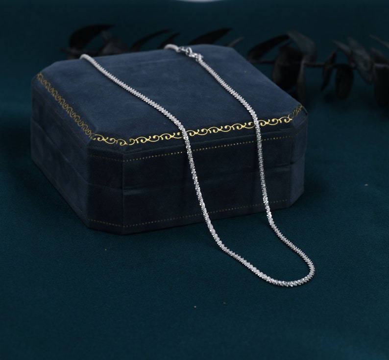 Puede incluir: Un collar de cadena de plata con peque&ntilde;as piedras transparentes engastadas en un dise&ntilde;o delicado. El collar se muestra en una caja de terciopelo azul.