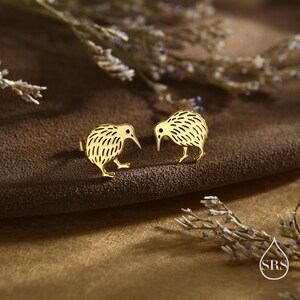 May include: A pair of gold-colored kiwi bird stud earrings. The earrings are detailed with etched lines and have a small post for securing them to the ear. The earrings are displayed on a brown surface.