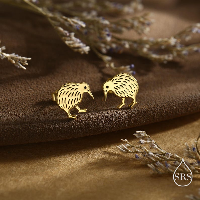 May include: A pair of gold-colored kiwi bird stud earrings. The earrings are detailed with etched lines and have a small post for securing them to the ear. The earrings are displayed on a brown surface.