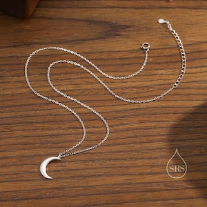 May include: A silver crescent moon pendant necklace on a delicate silver chain. The necklace is displayed on a wooden surface.