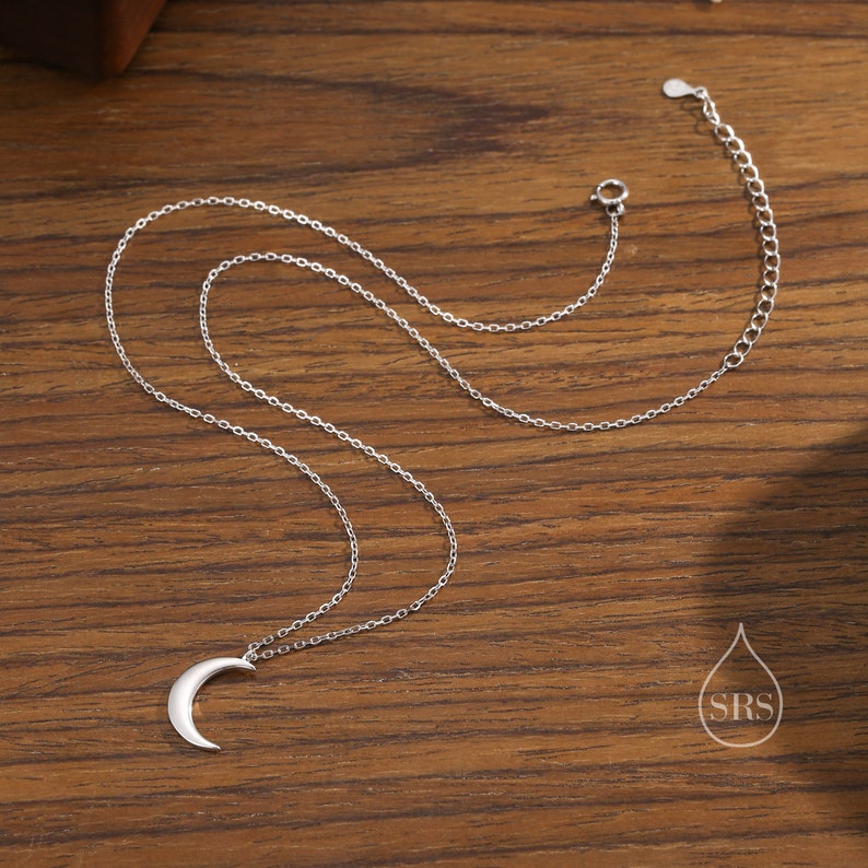 May include: A silver crescent moon pendant necklace on a delicate silver chain. The necklace is displayed on a wooden surface.
