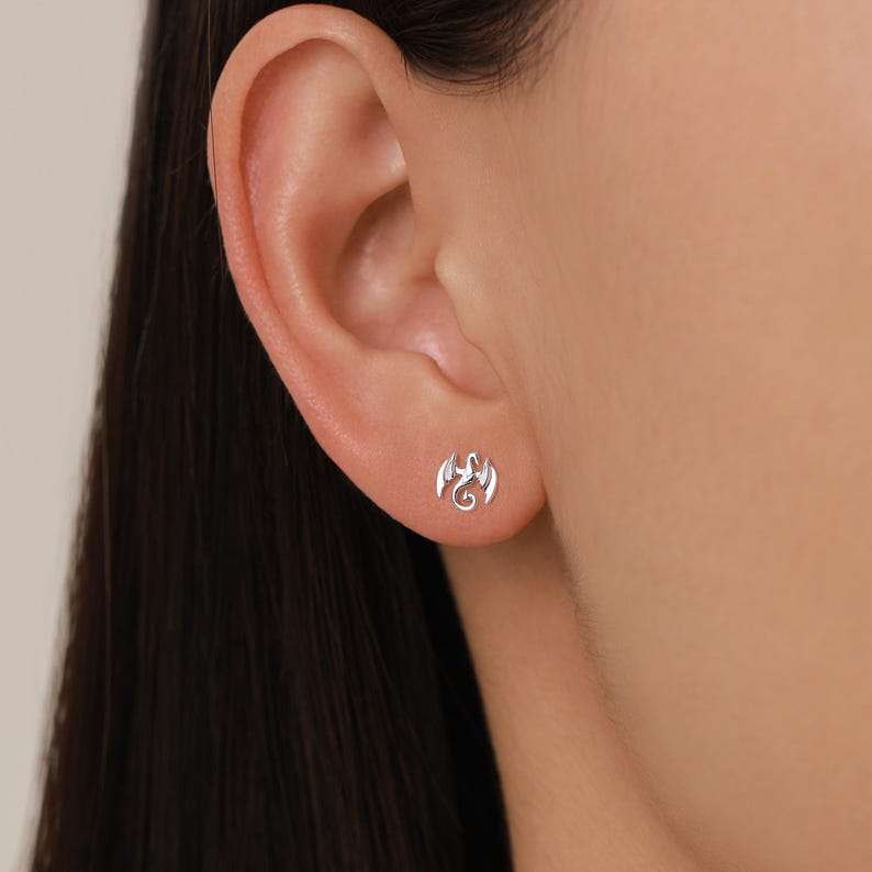 Peut inclure: Une boucle d'oreille en forme de dragon argent&eacute; est montr&eacute;e sur une oreille. La boucle d'oreille est une petite repr&eacute;sentation d&eacute;taill&eacute;e d'un dragon. C'est un bijou simple et &eacute;l&eacute;gant.