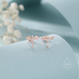 K&ouml;nnte beinhalten: Ein Paar ros&eacute;goldene Drachen-Ohrstecker. Jeder Ohrring zeigt einen detaillierten Drachen mit ausgebreiteten Fl&uuml;geln und einem gebogenen Schwanz. Die Ohrringe werden auf einer hellblauen Oberfl&auml;che pr&auml;sentiert.