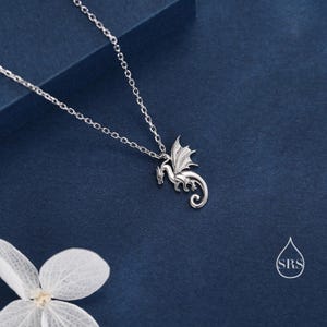 May include: A silver-toned necklace featuring a delicate dragon pendant with detailed wings and a curled tail. The pendant hangs from a thin, matching silver-toned chain. A small white watermark logo with the letters SRS is visible in the corner.