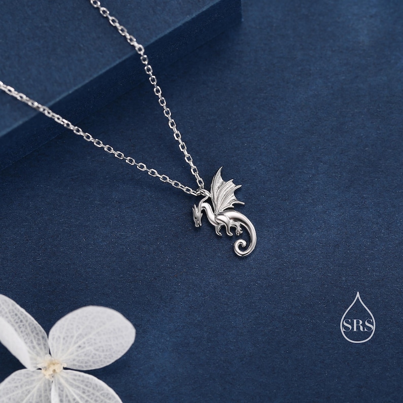 May include: A silver-toned necklace featuring a delicate dragon pendant with detailed wings and a curled tail. The pendant hangs from a thin, matching silver-toned chain. A small white watermark logo with the letters SRS is visible in the corner.