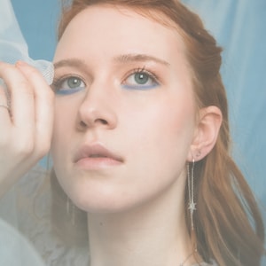 May include: A woman with red hair wearing a light blue dress and silver star earrings. She is holding a sheer white fabric in front of her face.