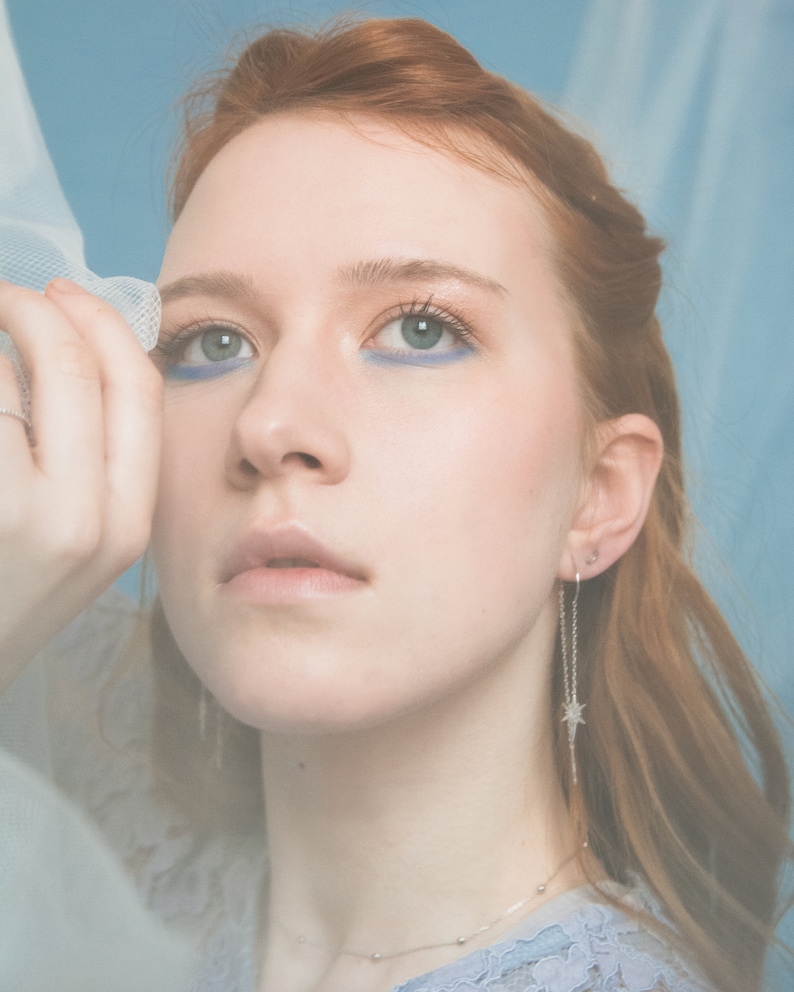 May include: A woman with red hair wearing a light blue dress and silver star earrings. She is holding a sheer white fabric in front of her face.