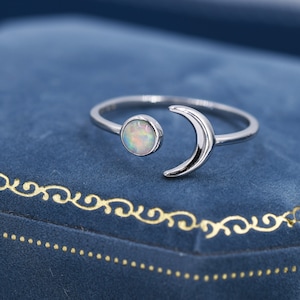May include: A silver ring featuring a moon and opal design. The ring has a crescent moon shape and a round opal stone. The opal has a mix of colors. The ring is set against a blue background.