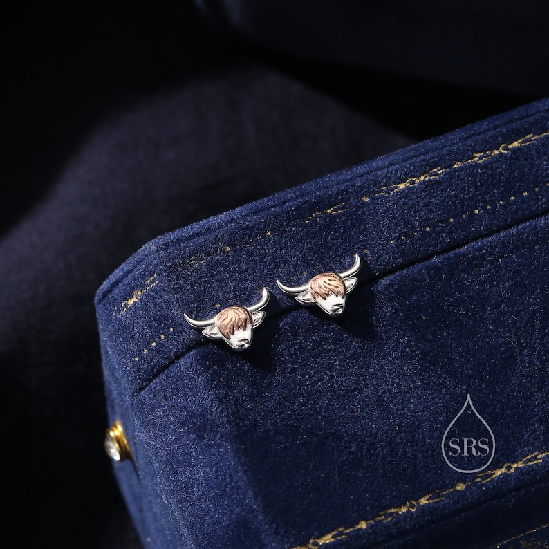 May include: A pair of silver earrings with a Highland cow design. The earrings are on a blue velvet background.