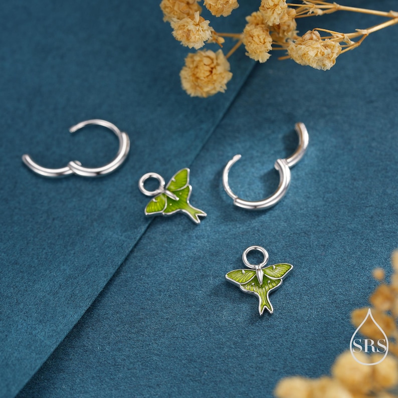 May include: Silver hoop earrings with green and silver moth charms. The earrings are set against a teal background with dried flowers. The moth charms have green enamel wings and detailed silver bodies.