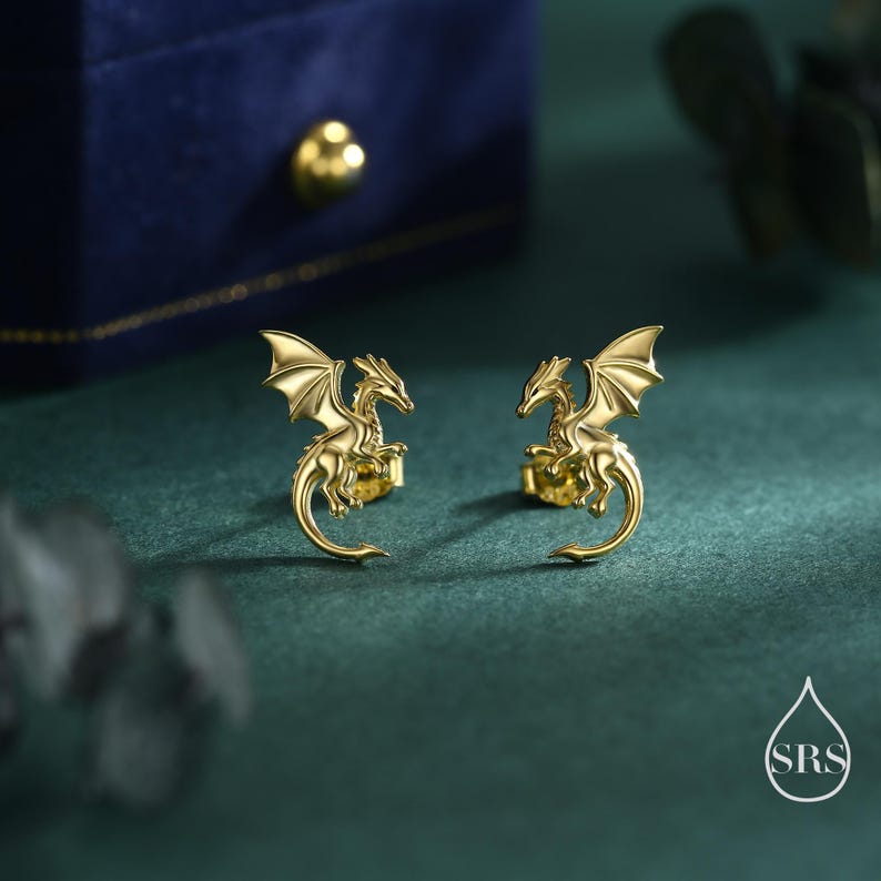 May include: A pair of gold dragon stud earrings. Each earring depicts a detailed dragon with outstretched wings and a curved tail. The earrings are presented on a dark green surface with a blue jewellery box in the background.