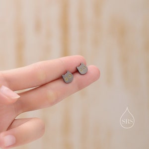 Peut inclure: Une paire de boucles d'oreilles clous en forme de chat noir avec des accents dor&eacute;s. Les boucles d'oreilles sont montr&eacute;es sur un doigt.
